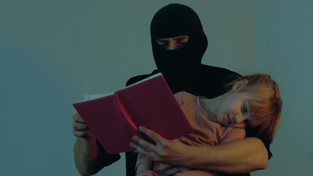 Kidnapper in a black balaclava holding a child in his arms, using book and reading. Little girl sleeping. Copy space. Child abduction concept.