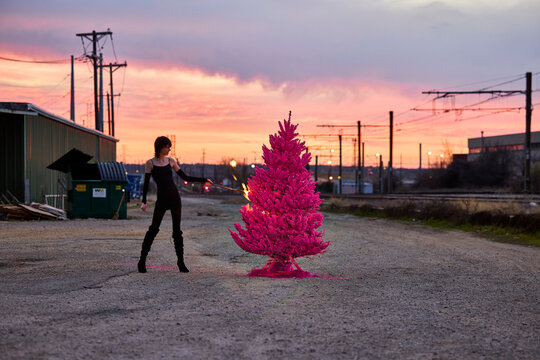Woman stands beside bright pink Christmas tree in urban industrial lot at sunset