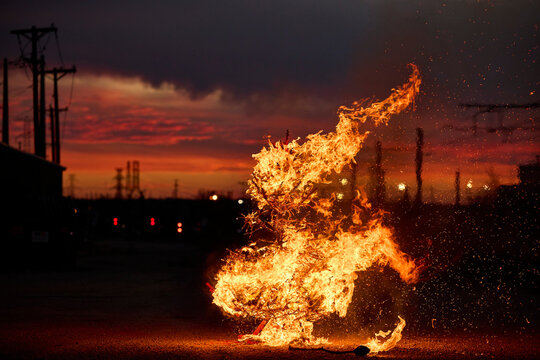 Dramatic flames and sparks illuminate an industrial landscape at sunset with power lines silhouetted