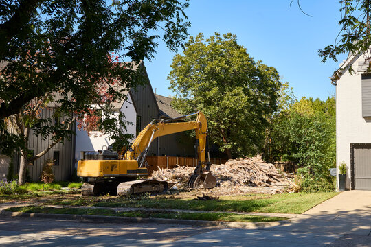 Yellow excavator demolishing residential property with debris pile in suburban neighborhood on sunny day