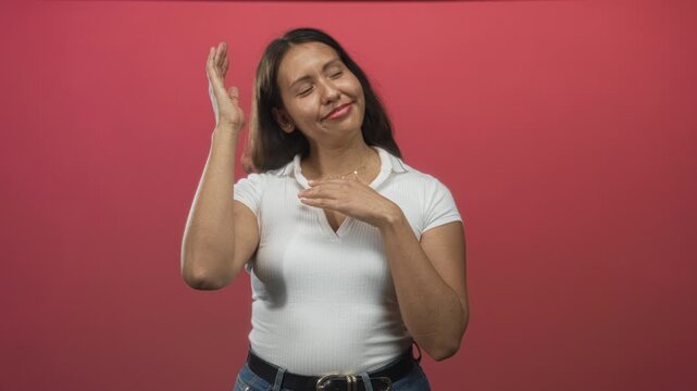 Woman raising right hand and flat left hand under chin wearing white polo and belt in pink studio with denim jeans; playful joy.
