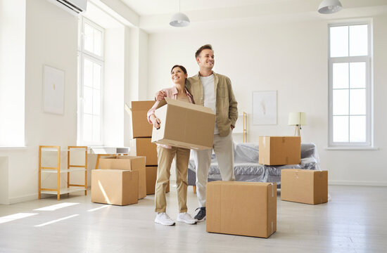 Happy couple moving cardboard boxes into new apartment. Sunlit living room with big windows, minimal furniture and scattered packing gear. Relocation, new beginnings at home and real estate