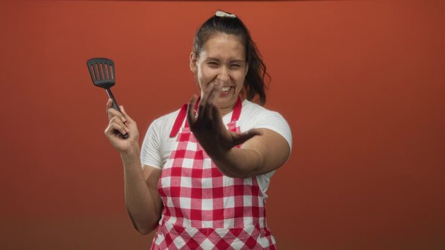 Woman cook holding spatula pointing finger toward camera in orange studio; home cooking confidence kitchen.