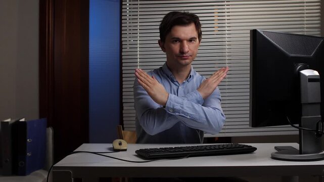 Young businessman sitting at his office desk crossing his arms in a firm no gesture, expressing refusal, boundary setting, or objection in a workplace setting