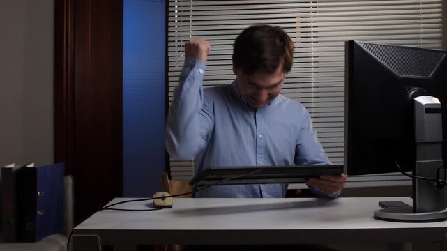 Young businessman experiencing extreme office stress, expressing rage and frustration by hitting his computer keyboard at a desk in an office environment