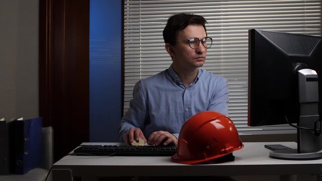 Young male engineer wearing glasses works at a computer in an office, focused on design and planning with an orange hard hat beside him and blueprints on the desk