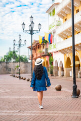 Unrecognized young Latina woman walks peacefully down a colonial street in Cartagena, Colombia, wearing a blue dress, white shoes, and a straw hat, surrounded by vibrant buildings and flags