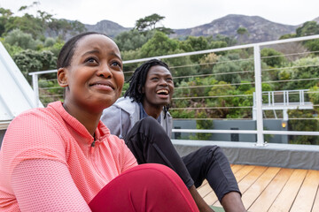 African American couple sitting on rooftop deck by cable railing on yoga mat in sportswear smiling