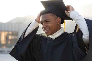 African American teen adjusting cap, smiling on campus in black gown white turtleneck gold tassel