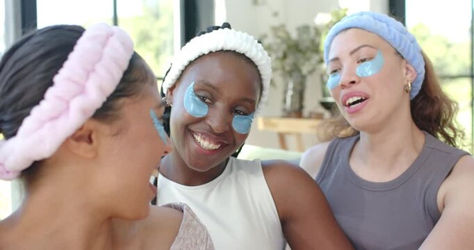 African American female friends wearing bands under-eye patches laughing talking on sofa for spa