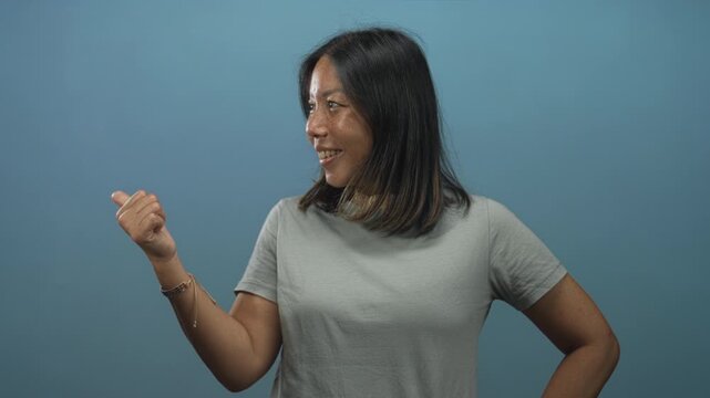 Woman with thumb raised giving a thumbs up gesture in a blue studio with gray t shirt and bracelet; approval friendliness.