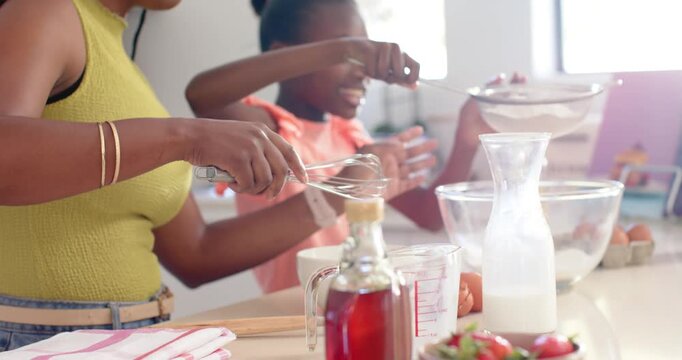 Mother whisking, guiding daughter tapping sieve, sifting flour into bowl for cake at kitchen island