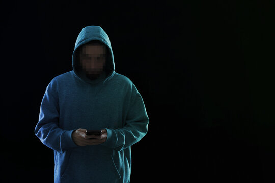 Criminal in hoodie using mobile phone on black background. Man with unrecognizable pixelated face