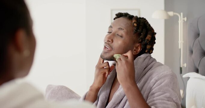 African American couple man raising guasha stone doing jawline massage checking skin in robe on bed