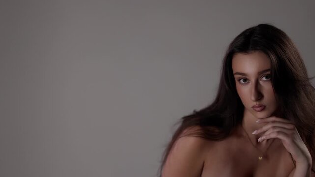 Slow motion beauty portrait of a sensual brunette model in a studio setting The model has perfect skin and is posing with her hand on her chin looking at the camera with a seductive gaze against a cle