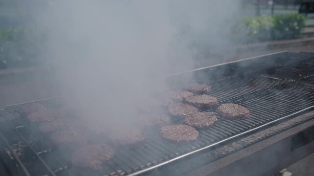 Burgers on greasy smoky charcoal BBQ on summer afternoon on commercial grill in slow motion