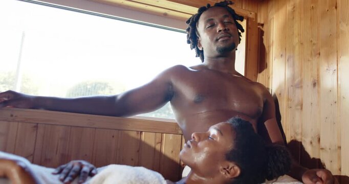 African couple seeking relaxation in spa, shirtless man glancing as woman breathing on rolled towel