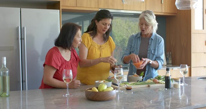 Diverse female friends gathering at kitchen island, preparing salad and adding carrot shreds