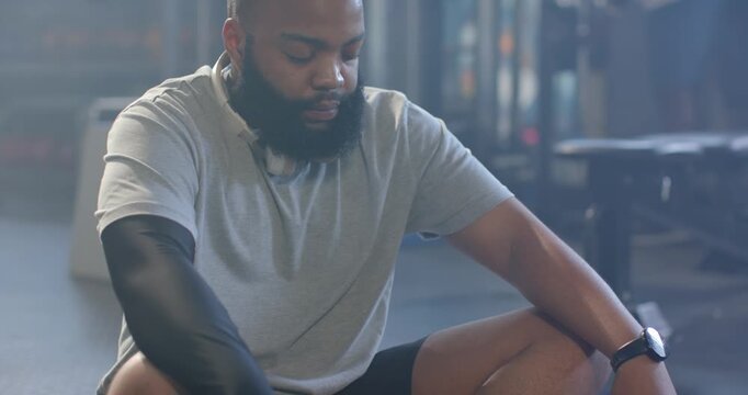 African male in sportswear sitting in gym adjusting arms, placing headphones, breathing to recover