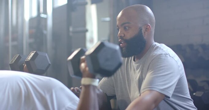 African American men trainer reaching spotting client pressing hex dumbbells on bench in gym safely