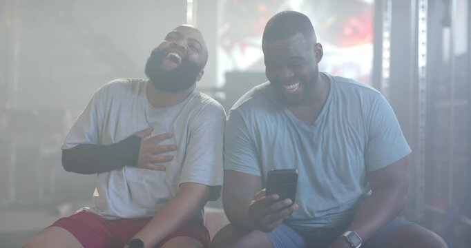 Two African men sitting at gym bench, right man showing funny smartphone clip, left man laughing