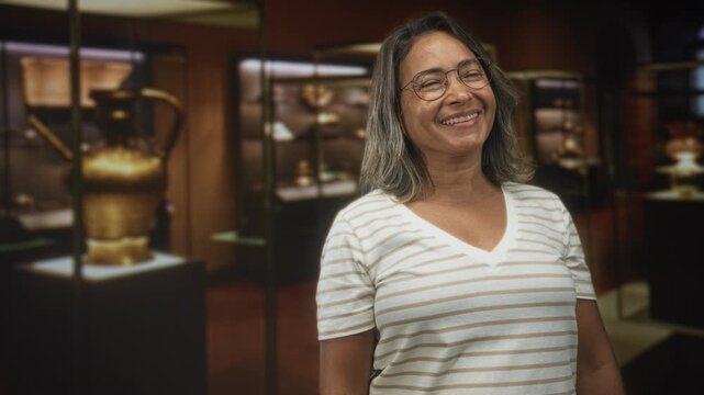 Hispanic woman smiling with closed eyes, head tilted, wearing glasses and a striped vneck shirt beside glass museum display cases with golden artifacts; contentment admiration.