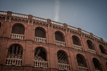 Naklejka premium Low angle view of Plaza de Toros de Valencia, a red brick bullring in Valencia, Spain, with arched galleries and white balustrades under a cloudy sky, highlighting a historic bullfighting landmark.