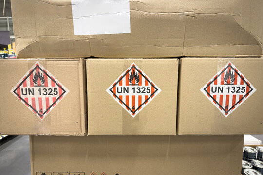 Cardboard packages stacked in a warehouse with UN 1325 hazard labels for flammable solids, class 4.1, showing flame warning symbols, dangerous goods logistics, safety compliance and shipping rules.