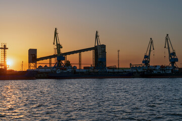 Obraz premium Port cranes for cargo transshipment on the mooring line of the Astrakhan port on the right bank of the Volga River against the background of the setting sun, Astrakhan, Russia