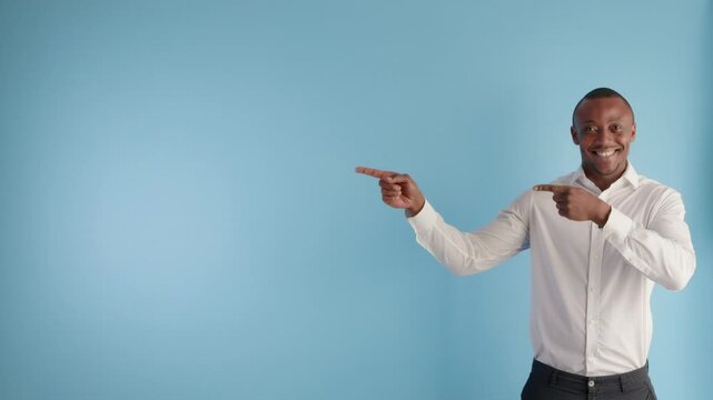 A smiling young Black man in a white shirt points with both index fingers to the empty blue background, offering space for text or graphics.