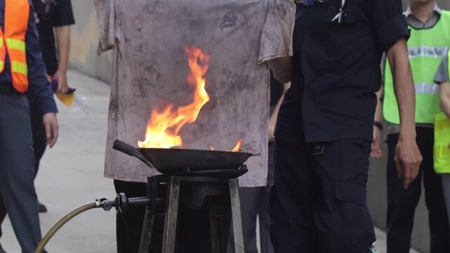 Fire safety training drill firefighter uses fire extinguisher on controlled flame wok oil pan, outdoor instructor led emergency practice