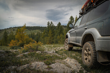 Off-road vehicle on rocky mountain trail in Mongolia, close up of SUV wheels on rough terrain, expedition car in autumn forest under cloudy sky, adventure travel and 4x4 concept © ANDREY