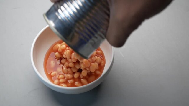 Close up of person pouring baked beans from tin can into white ceramic bowl on gray surface