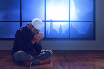 Young asian muslim man with beard praying near window at home, eid adha mubarak concept
