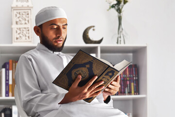 Young asian muslim man with beard reading holy book quran at home, eid adha mubarak concept