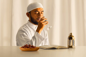 Young asian muslim man with beard drinking clear glass with water at home, eid adha mubarak concept