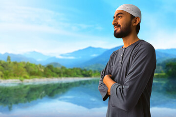 Portrait of young asian muslim man with beard standing and smiling on beautiful lake, eid adha mubarak concept