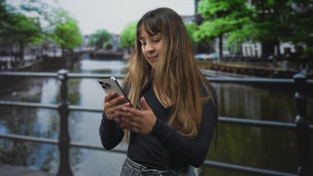 Young woman holding smartphone and texting by canal railing on a street, long hair, casual sweater and jeans showing a turning grimace into a smile; amused curiosity.