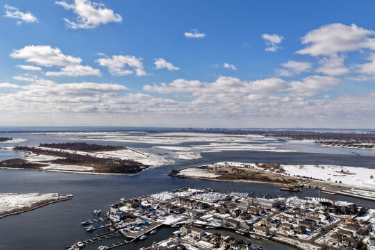 Oceanside, new york, showing an aerial panoramic view of a residential winter landscape with snow covered terrain, a marina with boats, and partially frozen waterways under a blue sky