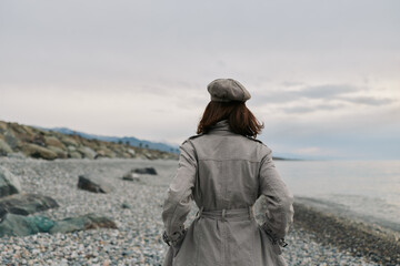 Woman wearing coat and beret stands on rocky beach looking at cloudy sea horizon. Concept of autumn fashion, solitude, and peaceful outdoor lifestyle near water. © SHOTPRIME STUDIO