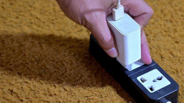 Hand unplugging white USB power adapter from black multi outlet power strip on brown carpet