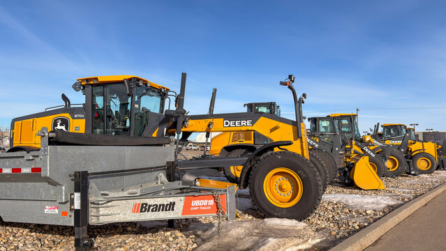 Regina, Canada - February 2026: Row of John Deere motor graders displayed at equipment dealership yard with Brandt dump trailer, highlighting heavy machinery for construction and roadwork.