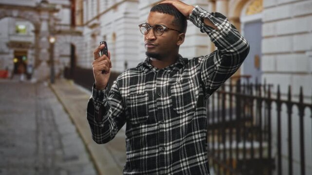 Man holding car keys with hand on head on street by building railings, wearing flannel shirt and glasses; uncertainty.