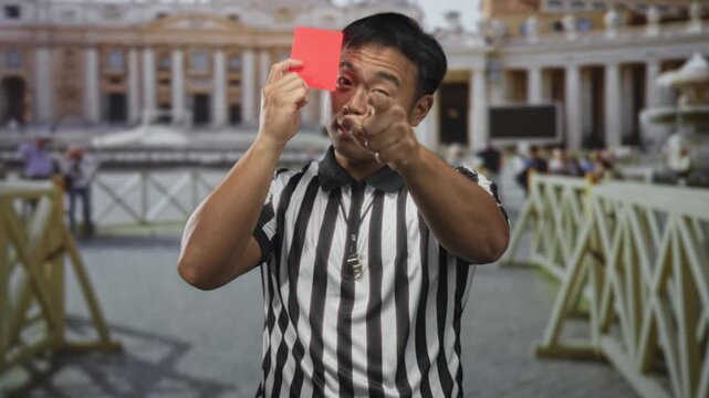 Man in referee uniform holds red card while blowing whistle and pointing finger in building; discipline.