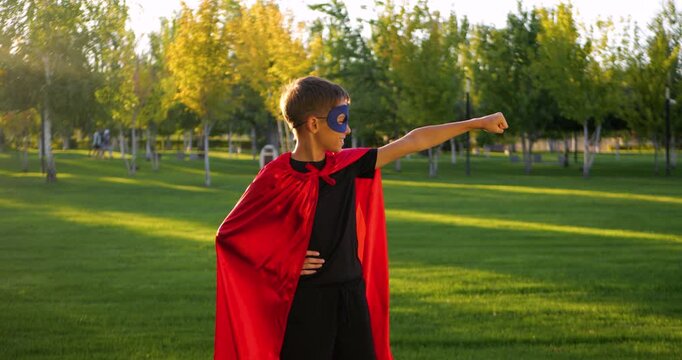 Child boy in red superhero cape stands in park. Kid wears blue mask and shows fist gesture. Child plays superhero game outdoors. Sunset light shines brightly. Costume party concept. Happy child.