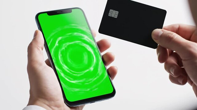 Close-up view of a hand holding a smartphone and a black credit card with green screen display, illustrating mobile payment in a studio.