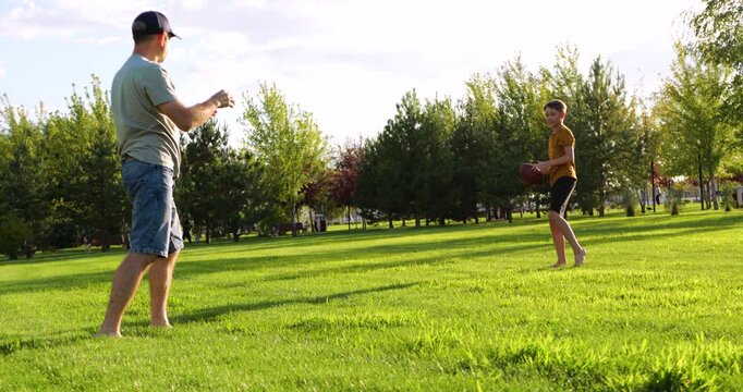 Dad and kid are playing rugby outside. Man throws ball while playing catch. Boy waits for ball while playing on lawn. Sport activity unites family. Barefoot people have fun in nature.