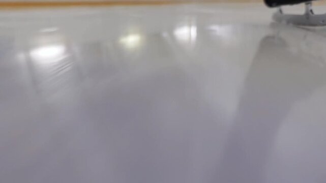 A dynamic close-up from ice height follows a figure skate blade in motion, blurring with speed against the frozen white surface. Indoor winter sports training.