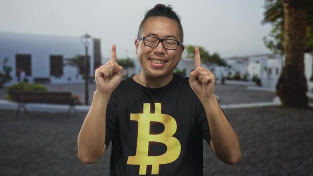 Man in bitcoin t shirt points both index fingers up on a street plaza showing hands and glasses with a broad smile; crypto optimism.