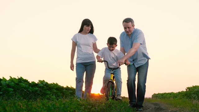 Father grips handlebar of bicycle while son pedals alongside mother on country road. Mother places hand on son acquiring skill of bicycle riding with assistance of father at twilight. Family vacation